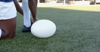 Çimlerin üzerinde diz çökmüş, rugby Afrikalı Amerikalı erkek oyuncu beyaz rugby topunu tekmelemeye hazırlanıyor, kopyalama uzayı. Spor, atlet, tekme, hazırlık, saha, yarışma