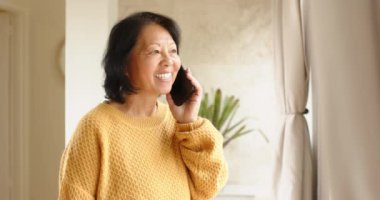 Sarı kazaklı yaşlı Asyalı kadın akıllı telefondan konuşuyor, pencerenin önünde gülümsüyor. Teknoloji, iletişim, mutluluk, yaşam tarzı, gündelik, kapalı alan