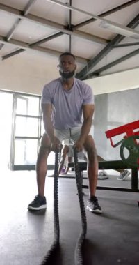 Dikey video: Adam spor salonunda savaş halatlarıyla egzersiz yapıyor, güç ve dayanıklılık kazanıyor. Fitness, antrenman, spor, kas, güç, atlet