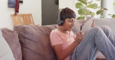 Kulaklık takan, kanepeye oturup tablet kullanan ve gülümseyen bir çocuk. Teknoloji, eğlence, eğlence, alet, rahatlama, çocukluk.