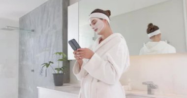 Yüz maskesi takmak, bornozlu bir kadın banyodaki spa rutininde akıllı telefon kullanıyor. Cilt bakımı, rahatlama, kendine bakım, sağlık, güzellik, şımartma.