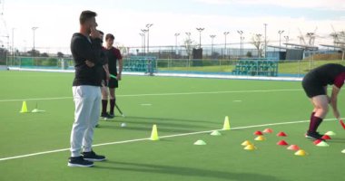 Koç, erkek hokey oyuncularını sahada külahlarla tatbikat yaparken gözlemliyor. Spor, antrenman, takım çalışması, koçluk, antrenman, atletizm
