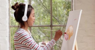 Evde müzik dinlerken tuvalde resim yapan Asyalı genç bir kadın. Sanat, yaratıcılık, hobi, kulaklık, ifade, eğlence