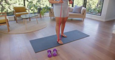 Direniş bandıyla egzersiz, artı oturma odasında yoga minderinin üzerinde duran boyunda bir kadın. Fitness, egzersiz, ev çalışması, direnç eğitimi, sağlık, sağlıklı yaşam tarzı.