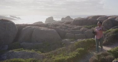 Kayalık sahil şeridinde yürüyen olgun Asyalı bir kadın elinde havluyla doğanın tadını çıkarıyor. Rahatlama, sağIık, sükunet, açık hava, manzara, keşif