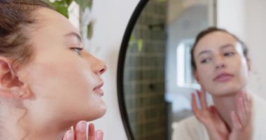 Aynaya bakan, yüzüne dokunan ve banyodaki deriyi inceleyen bir kadın. Cilt bakımı, kendine bakım, yansıma, güzellik, hijyen, bakım
