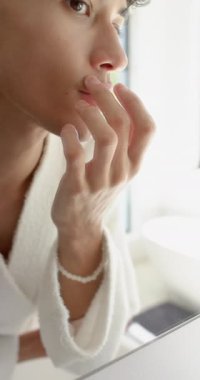 Dikey video: Bornozlu kişi banyo aynasında cildi inceliyor, özbakıma odaklanıyor. Cilt bakımı, kendini muayene, hijyen, bakım, sağlık, ev.