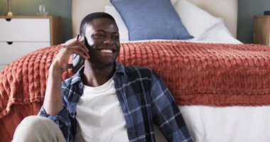 Afro-Amerikalı bir adam yatakta oturmuş, akıllı telefonuyla konuşurken gülümsüyor. İletişim, teknoloji, yaşam tarzı, neşeli, sohbet, rahatlama