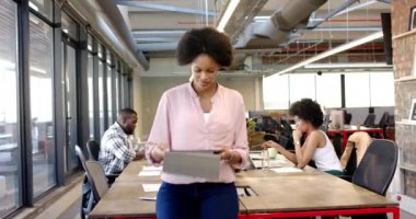 Afro-Amerikalı kadın modern ofiste kendinden emin bir şekilde tablet tutuyor. İş kurma gelişigüzel girişimci değişmemiş iş arkadaşları parlak, açık çalışma alanlarında dizüstü bilgisayarlarla masalarda çalışıyorlar.