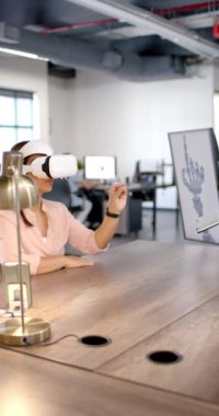 Dikey video: VR kulaklık kullanan kadın, ofiste bilgisayar ekranında robotik elle etkileşimde bulunuyor. Teknoloji, yenilik, sanal gerçeklik, robotik, etkileşim, işyeri