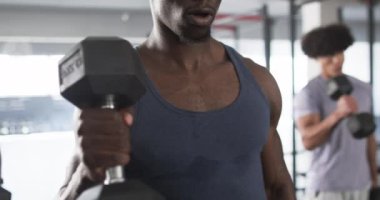 Spor salonunda mavi kolsuz tişörtlü, ağırlıklarla antrenman yapan genç bir Afrikalı Amerikalı. Güçlü ve formda, kısa siyah saçlı, odaklanmış ifade, yoğun, değişmemiş, yavaş çekim