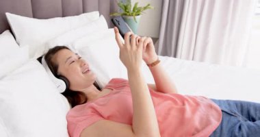 Yatakta uzanan, akıllı telefon kullanırken gülümseyen ve kulaklık takan bir kadın. Teknoloji, rahatlama, iletişim, dijital, yaşam tarzı, yatak odası