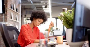 Modern ofisteki masasında çalışan kırmızı bluzlu melez bir kadın. İş kurma gelişigüzel girişimci değiştirilmemiş parlak, açık çalışma alanı bitkiler ve büyük pencereli, yavaş çekim