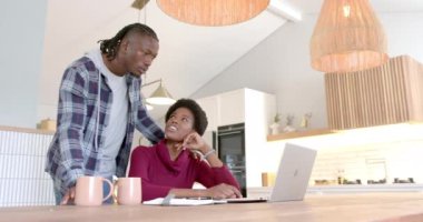 Evde çalışan Afro-Amerikan çift dizüstü bilgisayar kullanıyor ve mutfakta proje tartışıyorlar. Uzaktan çalışma, ev ofisi, takım çalışması, işbirliği, teknoloji, çalışma alanı