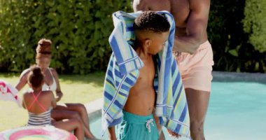 Havluyla kuruyan Afrikalı Amerikalı baba, havuzda yüzdükten sonra oğluna yardım ediyor. Afro-Amerikan ailesi, bağlılık, havuz başı, babalık, çocukluk, yaz