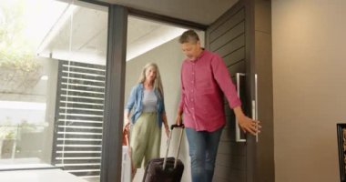 Eve gelen, bavulları gülen ve işaret eden çok ırklı yaşlı çift, kopyalama alanı. seyahat, varış, tatil, mutluluk, keşif, kapalı alan