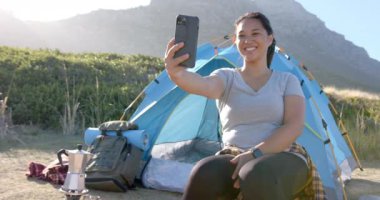 Selfie çeken, dağ yürüyüşü sırasında çadırın önünde oturan bir kadın. Macera, kamp, yürüyüş, seyahat, keşif, manzara