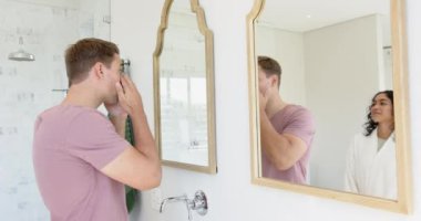 Genç, çok ırklı bir çift banyoda, erkek ellerini yıkarken Asyalı kadın aynaya bakıyor, evde. Hijyen, sabah rutini, ilişki, tımar, bağlanma, günlük yaşam