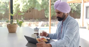 Kahvaltı yapıyor, türbanlı bir adam mutfak tezgahında otururken tablet kullanıyor. Teknoloji, sabah rutini, çoklu görev, yaşam tarzı, yerli, kolaylık