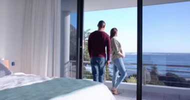 Kucaklayan, hamile bir çift okyanus manzaralı geniş pencerenin önünde dikiliyor. Aile, aşk, hamilelik, birliktelik, beklenti