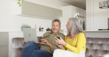 Kanepede oturmak, evde dizüstü bilgisayar ve akıllı telefon kullanan çok ırklı yaşlı bir çift. Teknoloji, yaşam tarzı, birliktelik, kapalı alan, çoklu görev, rahatlama