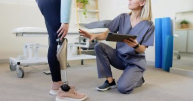 Sarışın, mavi gözlü, gri önlüklü beyaz protezci diz çökerken protez bacağını düzeltiyor. Bir tablet tutmak ve hasta ile uygun olup olmadığını tartışmak, değiştirilmemiş