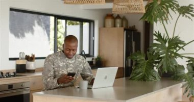 Modern mutfakta akıllı telefon ve dizüstü bilgisayar kullanarak askeri üniformalı gülümseyen adam. technology, communication, multitasking, domestic life, home, device