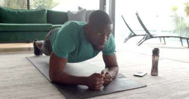 Güçlü ve formda Afro-Amerikan bir adam evde yoga minderi üzerinde kalas egzersizi yapıyor. Spor, spor, spor salonu, güç, dayanıklılık, değişmemiş, yaşam tarzı.