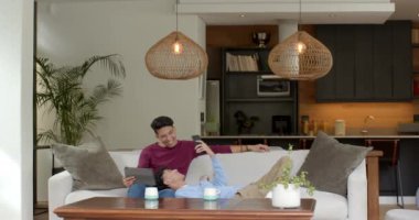 Kanepede dinlenen, çok ırklı eşcinsel çift evdeki rahat oturma odasında tabletin tadını çıkarıyor. Rahatlama, teknoloji, kapalı alan, samimiyet, birliktelik, rahatlık