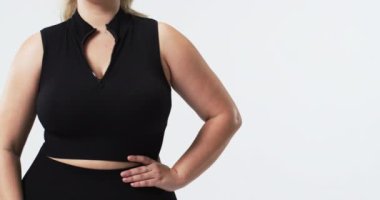 Beyaz, güzel, iri yarı bir kadın siyah kolsuz bluz ve uyumlu pantolonla poz veriyor. Düz beyaz arka plan vücudunun pozitifliğini ve kendine güvenini vurgular, değişmez.