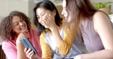 Üç bayan arkadaş akıllı telefona bakıyor, gülüyor ve evde birlikte vakit geçiriyorlar. Arkadaşlar, birliktelik, teknoloji, kapalı alan, boş zaman, sosyalleşme