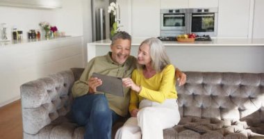 Kanepede oturan, modern ev mutfağında gülümseyen ve tablet kullanan çok kültürlü yaşlı bir çift. Teknoloji, mutluluk, yaşam tarzı, birliktelik, iletişim, rahatlama