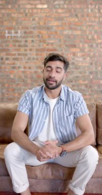 Dikey video: Adam kanepede oturmuş elleriyle el kol hareketi yapıyor, bir şeyleri gündelik bir ortamda açıklıyor. İletişim, tartışma, etkileşim, sohbet