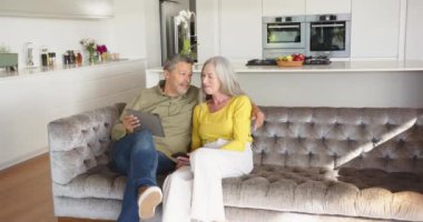 Kanepede dinlenmek, modern mutfaktaki tablet ve akıllı telefon kullanan çok ırklı yaşlı bir çift. Teknoloji, yaşam tarzı, rahatlama, birliktelik, bağlantı, gelişigüzel