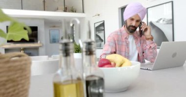 Telefonda konuşan, evde meyve kasesiyle otururken dizüstü bilgisayar kullanan bir adam. Telekomünikasyon, çoklu görev, üretkenlik, ev ofisi, teknoloji, uzaktan çalışma