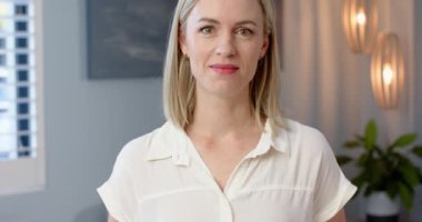 Beyaz, orta yaşlı, sarışın, mavi gözlü, kapalı alanda gülümseyen bir kadın. Beyaz bluz giyiyor, soyut resimler ve arka planda hafif ışıklandırma var, değişmemiş, yavaş çekim.