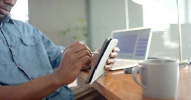 Tablette stilus kullanan Afro-Amerikan bir adam evde dizüstü bilgisayar ve kahveyle çalışıyor. Uzaktan çalışma, teknoloji, ev ofisi, kahve molası, çoklu görev, telekomünikasyon, değişmemiş, yaşam tarzı