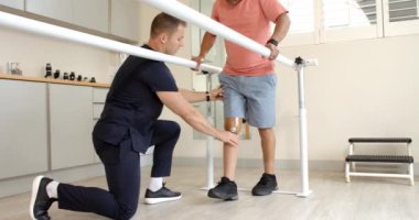 Klinikteki paralel barlarda yürüyen protez bacaklı bir adama yardım eden terapist. Fizyoterapi, rehabilitasyon, fiziksel, sağlık, tıbbi, destek