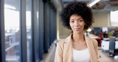 İş kıyafetleri içinde kollarını kavuşturarak güvenle gülümseyen melez genç bir kadın. Geniş pencereli ofis ortamı ve arka planda modern mobilyalar, değiştirilmemiş