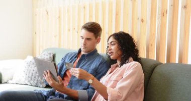 Genç, çok ırklı bir çift evdeki koltukta online alışveriş için tablet ve kredi kartı kullanıyor. ekommerce, yaşam tarzı, teknoloji, ev, dijital, ödeme