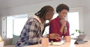 Gülümseyen Afrikalı Amerikalı çift ellerinde kahve fincanları ve tabletlerle not defterine yazıyorlar. Rahatlatıcı, birliktelik