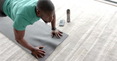 Yoga minderinde şınav çeken, evde su şişesiyle egzersiz yapan Afrikalı Amerikalı bir adam. Fitness, egzersiz, ev egzersizi, dayanıklılık eğitimi, su kaybı, değişmemiş, yaşam tarzı