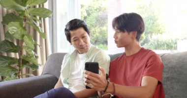 Asyalı baba ve genç oğul kanepede oturuyor, oğlu akıllı telefondan bir şey gösteriyor. Parlak oturma odası, geniş pencereli ve yemyeşil, değişmemiş.