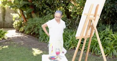 Tabloda resim yapan, bahçesinde sehpa ve boya malzemeleriyle duran yaşlı bir kadın. Sanat, yaratıcılık, bahçıvanlık, hobi, boş zaman, açık hava