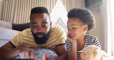 Baba ve kızı evde birlikte tablet kullanırken gülüyorlar. Afro-Amerikan ailesi, bağlanma, teknoloji, birliktelik, kapalı alan, ebeveynlik