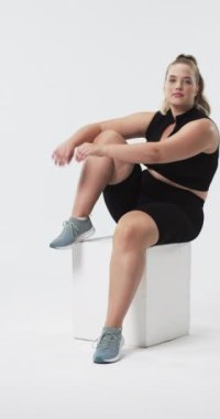 Dikey video: Atletik olarak gülümseyen ve beden pozitifliğini artırmak için beyaz küpün üzerinde oturan iri yarı bir kadın. Fitness, sağlık, güven, aktif giyim, güçlendirme, spor giyim