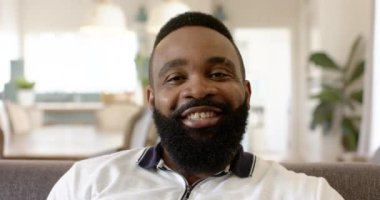 Evdeki koltukta oturan sakallı, gülümseyen adam kameraya bakıyor. Yaşam tarzı, konfor, rahatlama, mutluluk, portre, kapalı alan