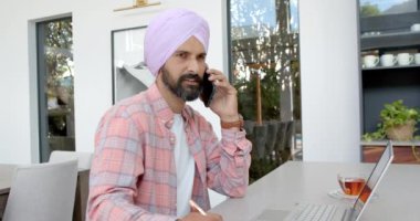 Akıllı telefondan konuşmak, evde dizüstü bilgisayar ve masa başında çayla çalışan bir adam. Uzaktan çalışma, telekomünikasyon, üretkenlik, teknoloji, ev ofisi, iş dünyası