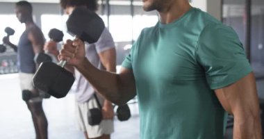 Spor salonunda antrenman yapan ve egzersizlerine odaklanan bir grup genç adam. Üçü de zinde, biri dambıl tutuyor, diğerleri ayakta, atletik kıyafetler giyiyor, değişmemiş, ağır çekim.