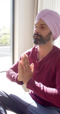 Dikey video: Evde meditasyon yapan, türbanlı bir adam oturma odasında farkındalık uyguluyor. Meditasyon, sağlık, rahatlama, huzur, sükunet, kişisel bakım.